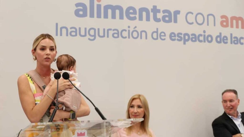 Woman at a Podium Holding a Baby and Speaking into Two Microphones, with a Seated Woman Nearby and a Spanish Backdrop Behind Them. - Entre Regios