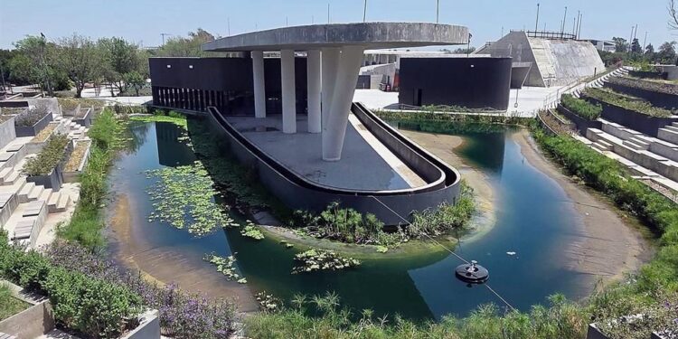 Modern Circular Pond with a Concrete Pavilion on a Curved Wall, Surrounded by Stepped Seating and Greenery in a Plaza. a Small Fountain Floats on the Water Surface. - Entre Regios